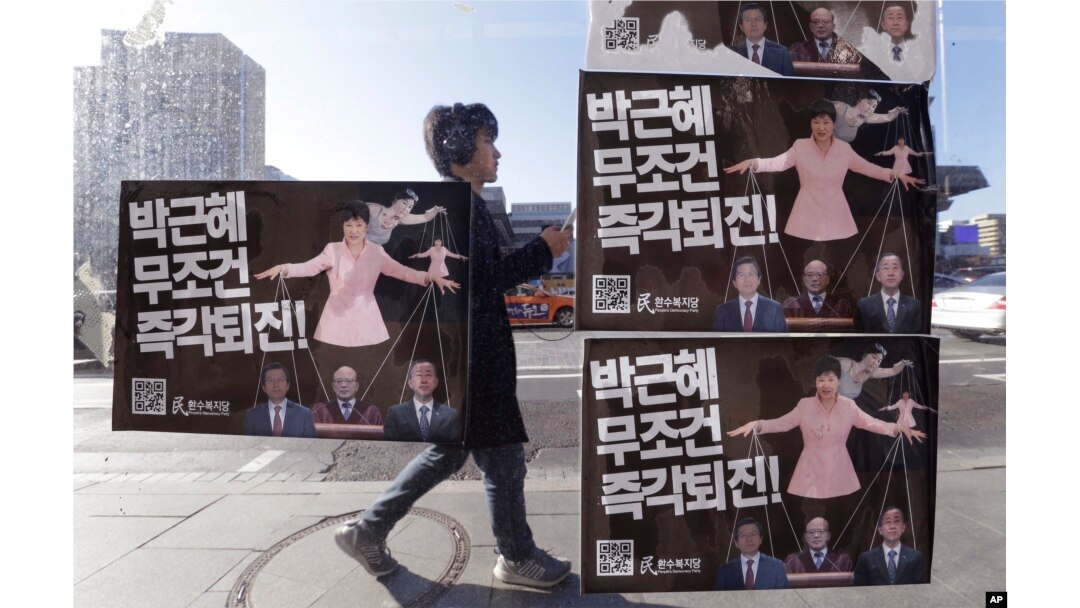 FILE - A man walks by a bus stop displayed with posters depicting impeached South Korea's President Park Geun-hye in pink manipulating three of her aides while she is also manipulated as a marionette by her jailed confidante Choi Soon-sil, seen above Park's left