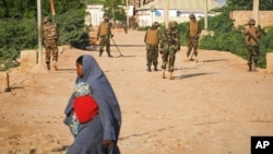 FILE - A Somali woman crosses a street while soldiers with the African Union Mission in Somalia (AMISOM) stand guard 300 kilometers west of the capital Mogadishu, on a street in Belet Weyne, Somalia. 