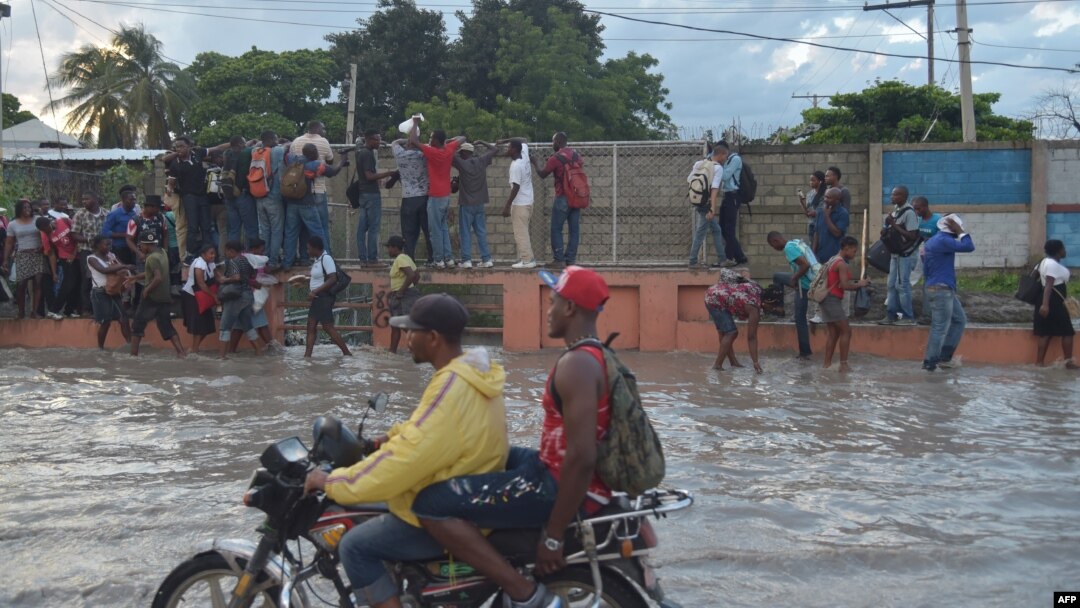 A group of people walk in a partially flooded street by the rains, while another persons try to walk over the water, holding a grating, in the Haitian capital Port-au-Prince, on May 2, 2017.