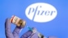 A woman holds a small bottle labeled with a "Coronavirus COVID-19 Vaccine" sticker and a medical syringe in front of displayed Pfizer logo in this illustration taken, Oct. 30, 2020.
